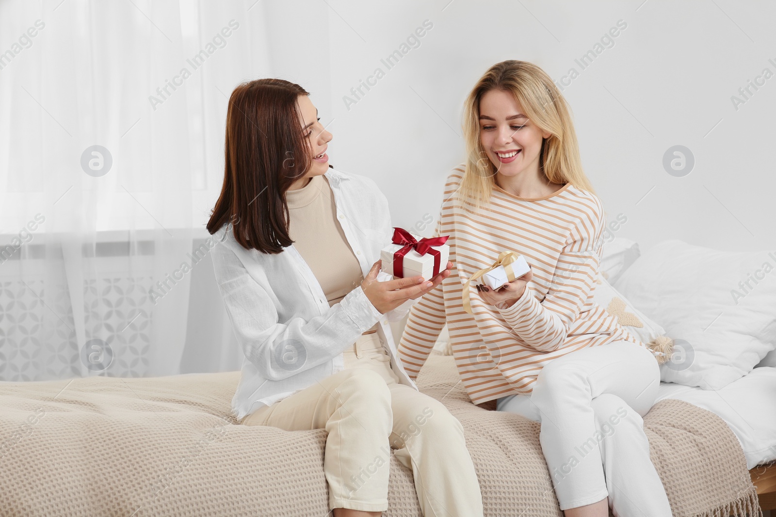 Photo of Smiling young women presenting gifts to each other on bed at home