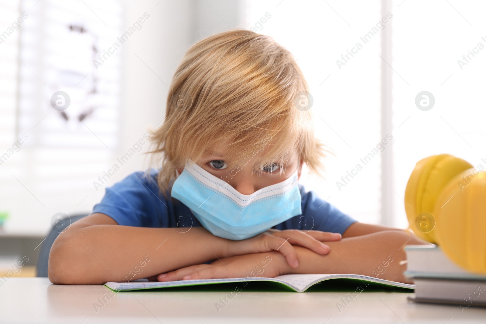 Photo of Sad little boy in mask with exercise book at home. Distance learning during COVID-19 pandemic