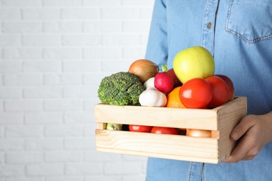 Photo of Woman holding wooden crate with fruits and vegetables at white brick wall, closeup. Space for text