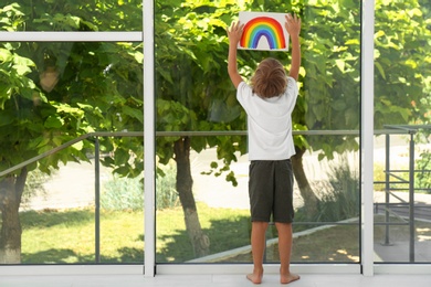 Photo of Little boy holding rainbow painting near window indoors. Stay at home concept