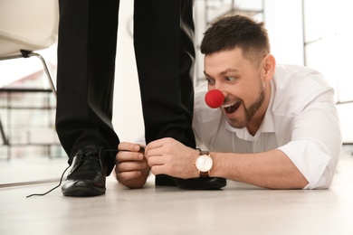 Man with clown nose tying shoe laces of his colleague together in office, closeup. Funny joke