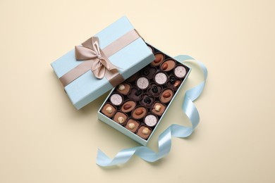 Photo of Open box of delicious chocolate candies and light blue ribbon on beige background, flat lay