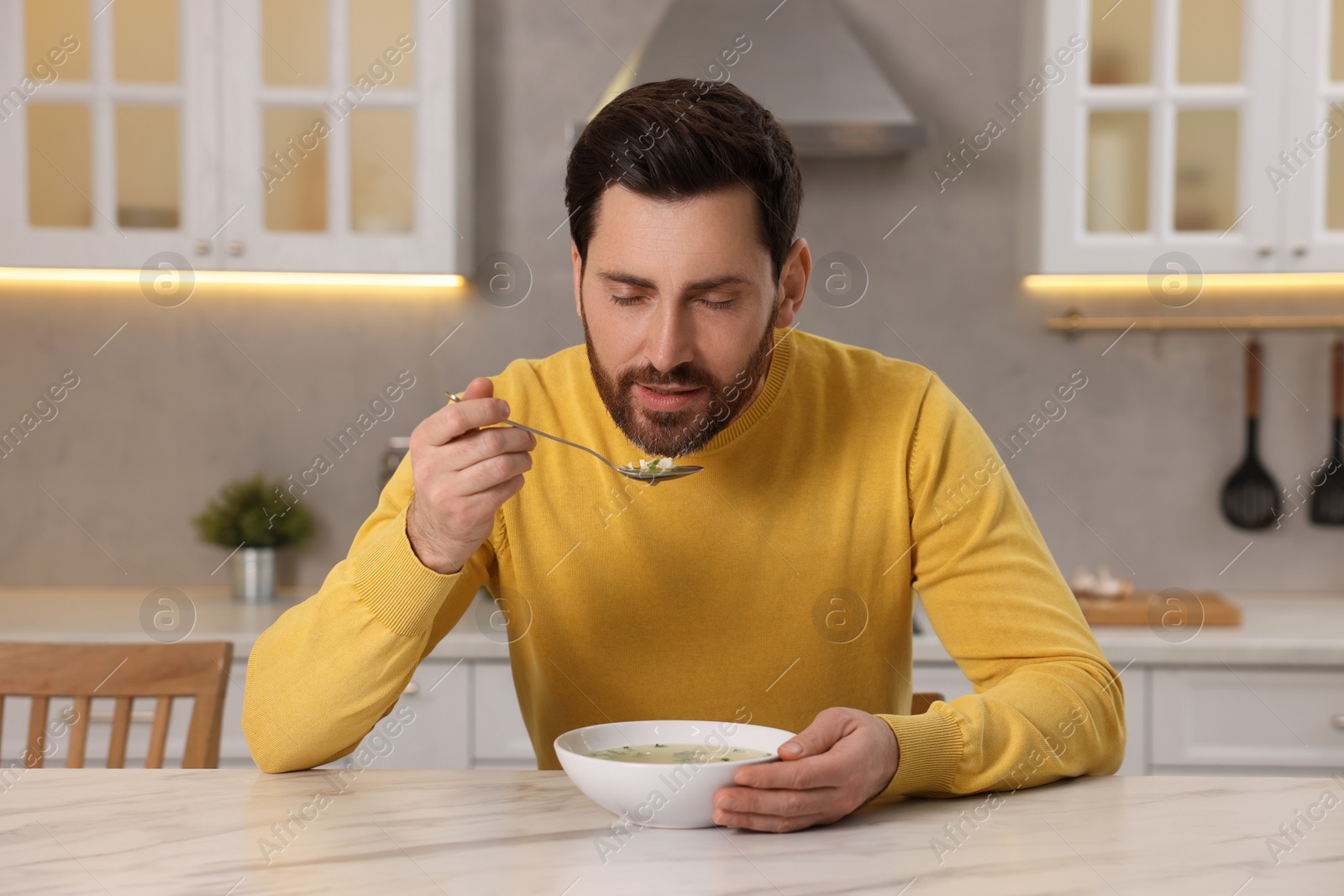 Photo of Man eating delicious chicken soup at light marble table in kitchen