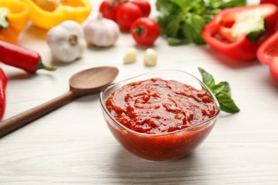 Delicious adjika sauce in glass bowl and ingredients on wooden table