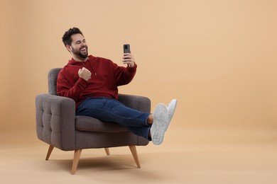 Photo of Happy young man using smartphone on armchair against beige background, space for text