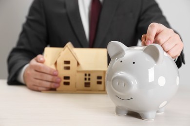 Man putting coin into piggy bank near house model at wooden table, closeup with space for text. Saving money concept