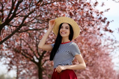 Pretty young woman with straw hat near beautiful blossoming trees outdoors. Stylish spring look