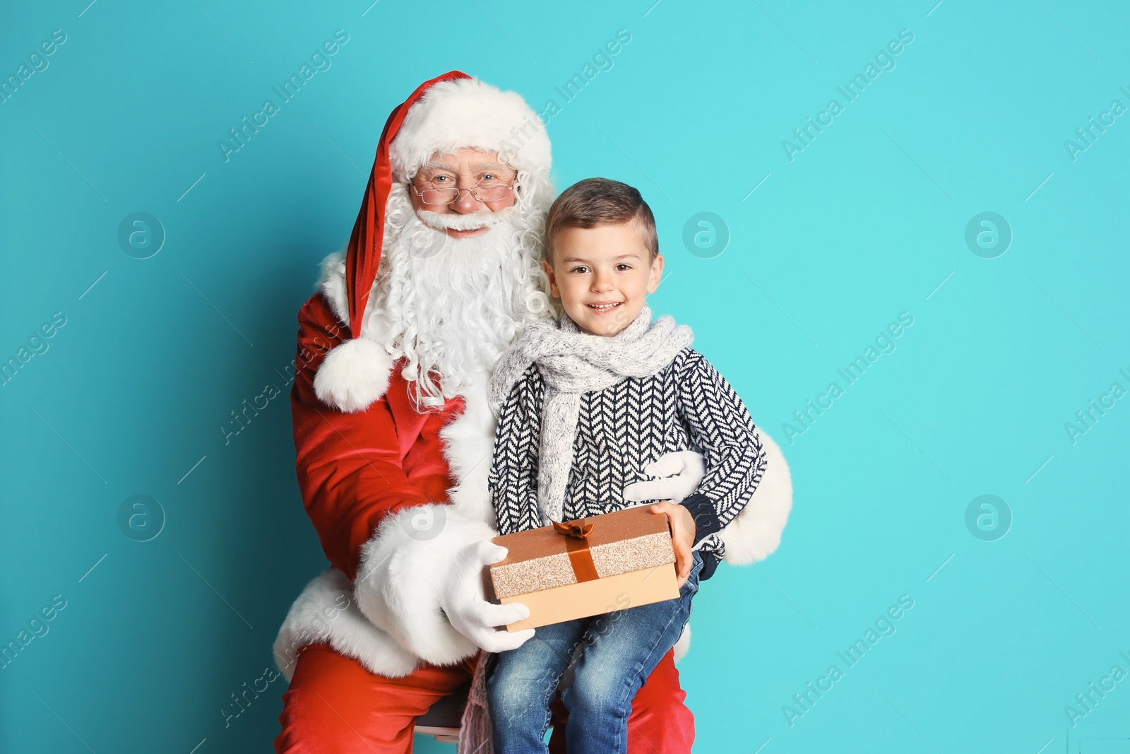 Photo of Little boy with gift box sitting on authentic Santa Claus' lap against color background