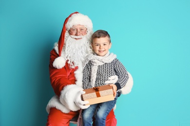 Photo of Little boy with gift box sitting on authentic Santa Claus' lap against color background