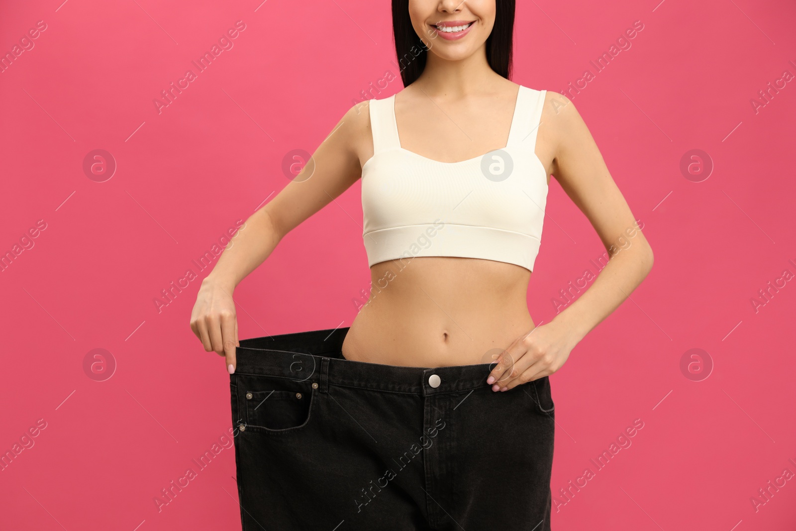 Photo of Happy young woman wearing big jeans after weight loss on pink background, closeup