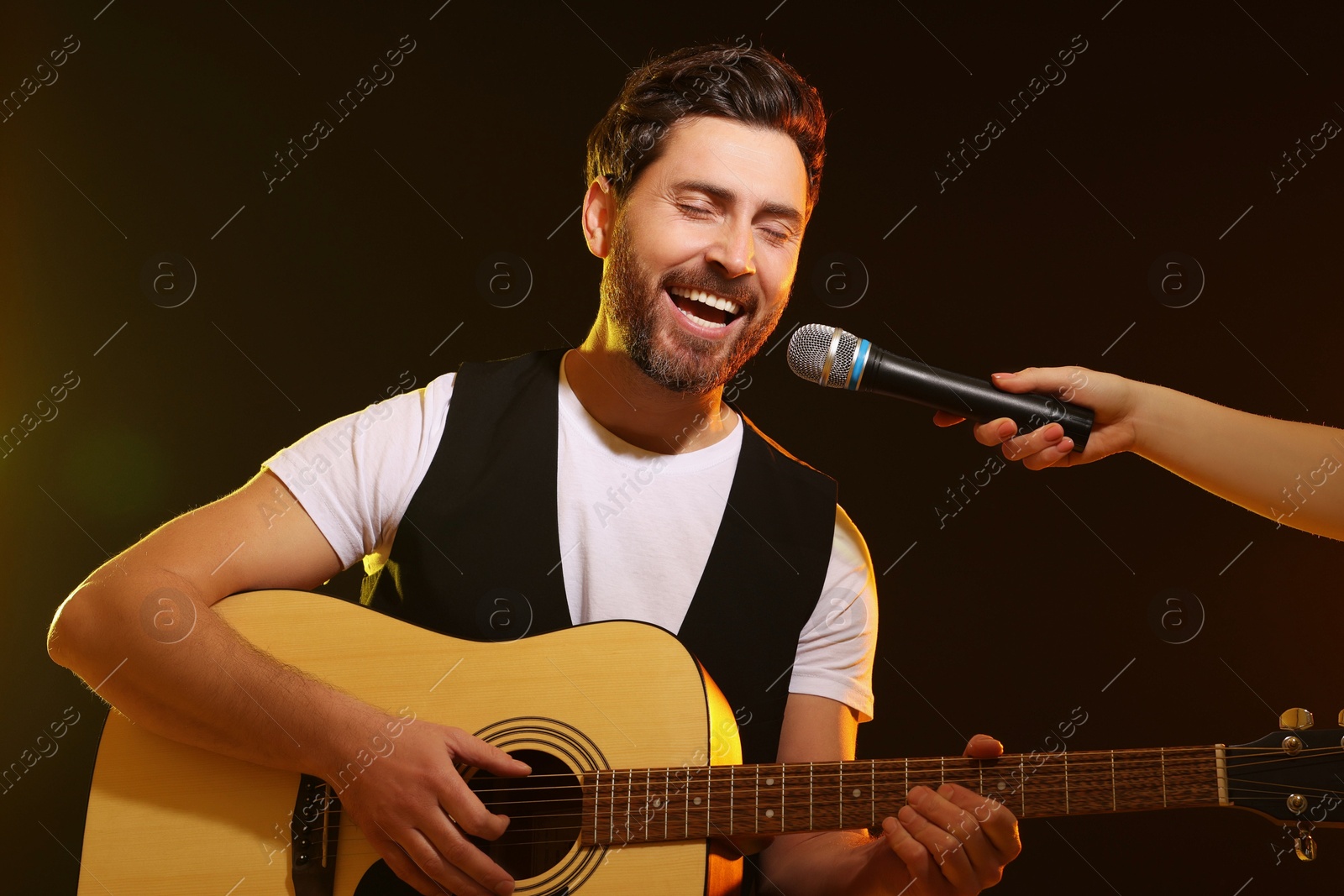 Photo of Handsome man with acoustic guitar singing while woman holding microphone on dark background, closeup