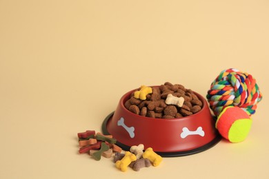 Photo of Dry pet food in bowl, vitamins and toys on beige background, space for text