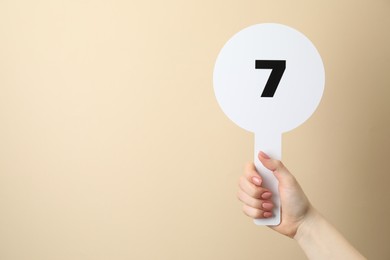Photo of Woman holding auction paddle with number 7 on beige background, closeup. Space for text
