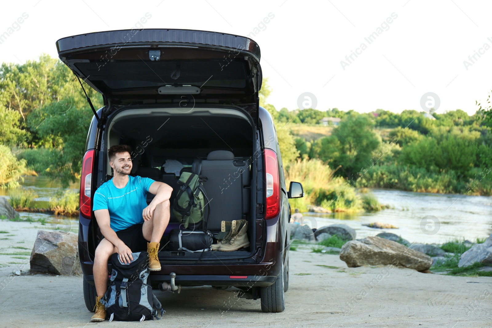 Photo of Young man near car with sleeping bag and camping equipment outdoors. Space for text