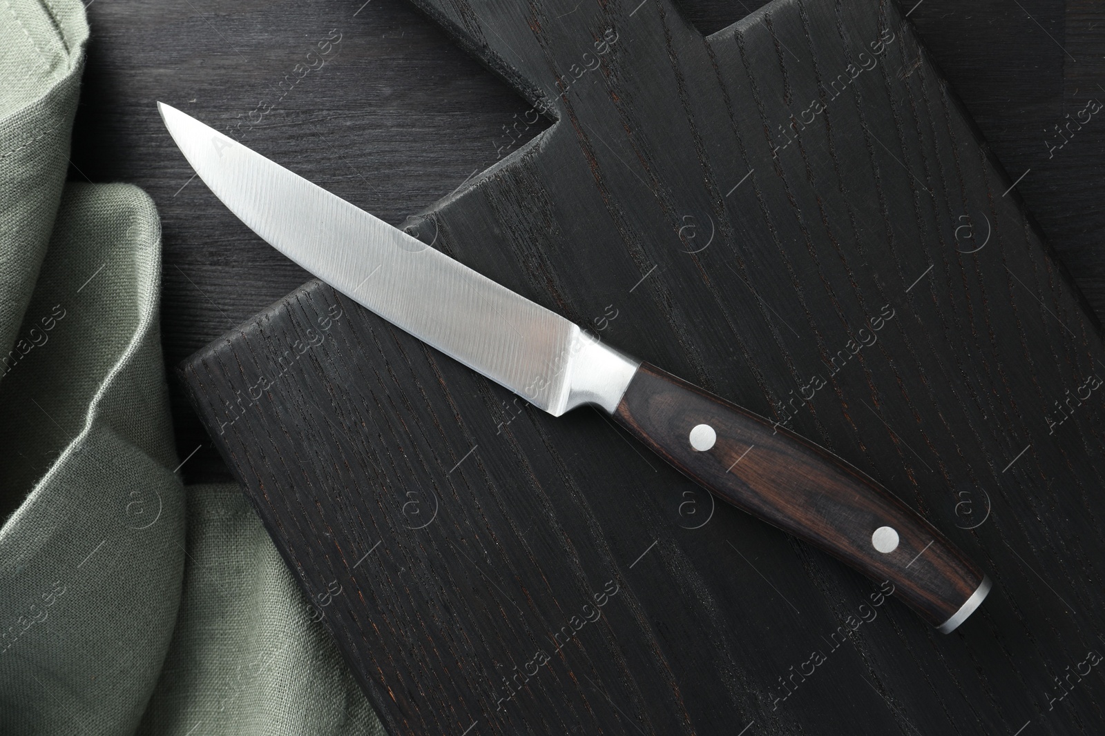 Photo of One sharp knife, cloth and board on black wooden table, top view