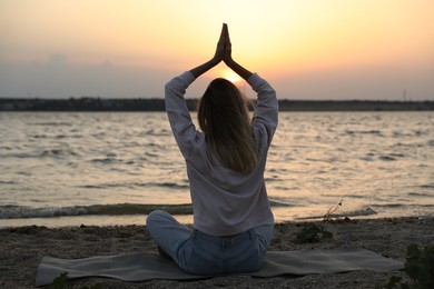 Photo of Young woman meditating on beach near river at sunset, back view