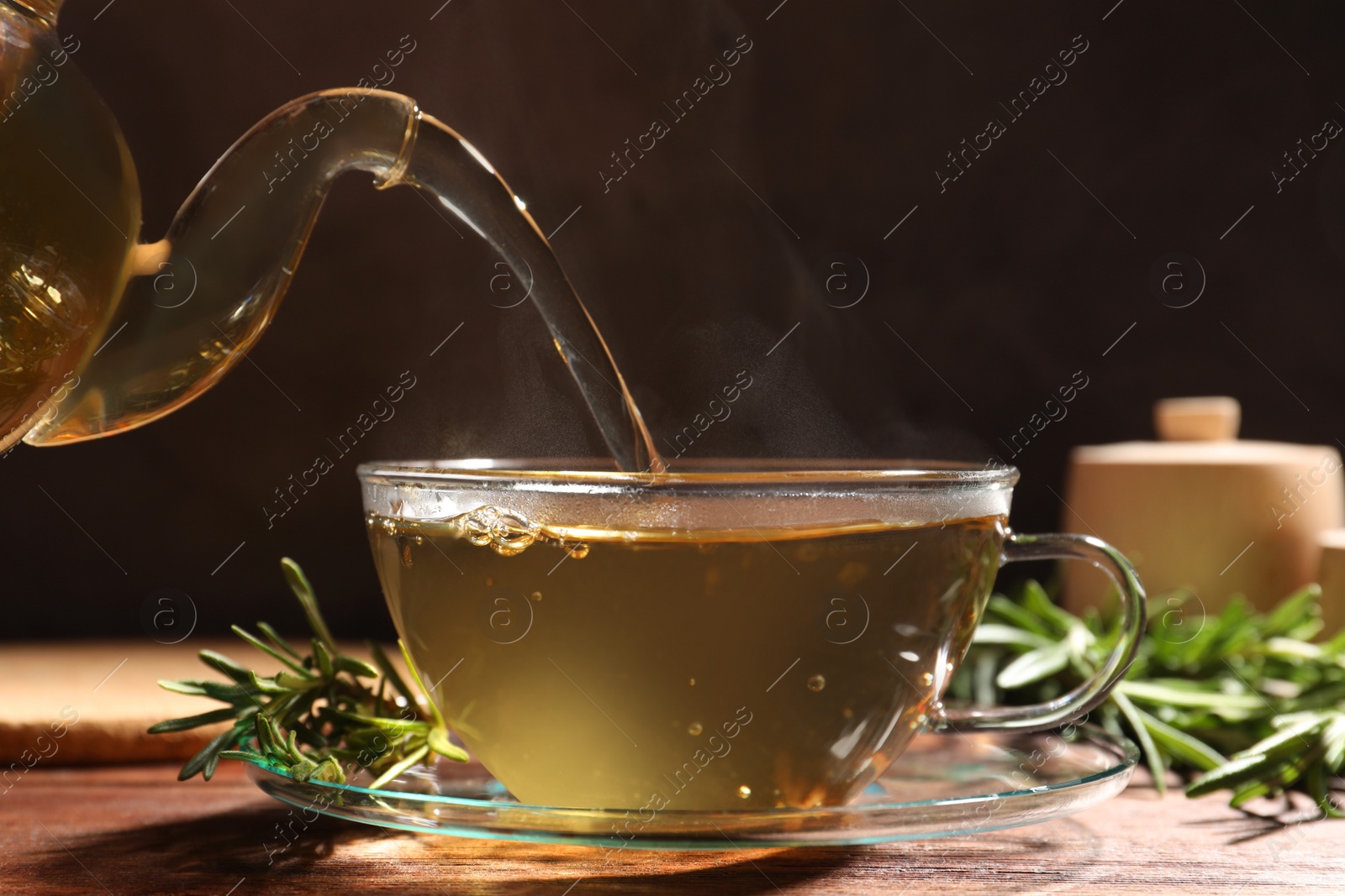 Photo of Pouring aromatic herbal tea into cup and fresh rosemary on wooden table