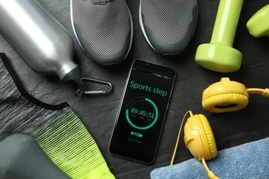 Smartphone with step counter app and fitness accessories on black background, flat lay