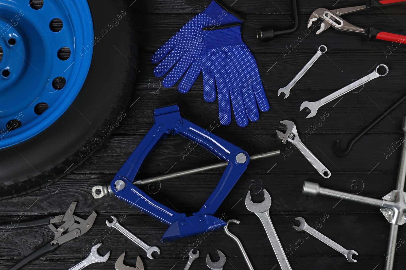 Photo of Car wheel, scissor jack, gloves and different tools on black wooden surface, flat lay