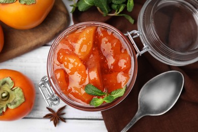 Jar of tasty persimmon jam and ingredients on white wooden table, flat lay