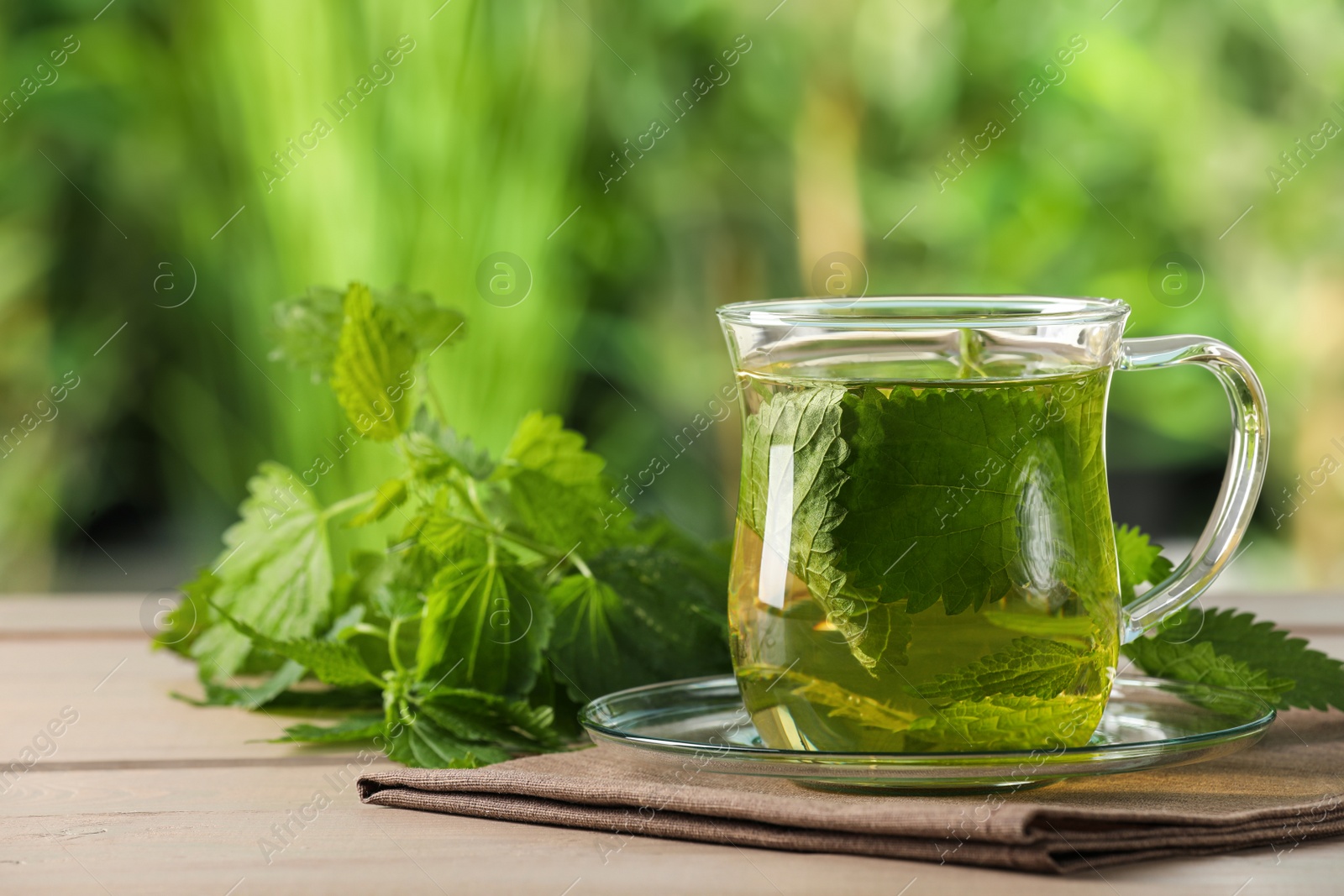 Photo of Glass cup of aromatic nettle tea and green leaves on wooden table outdoors, space for text