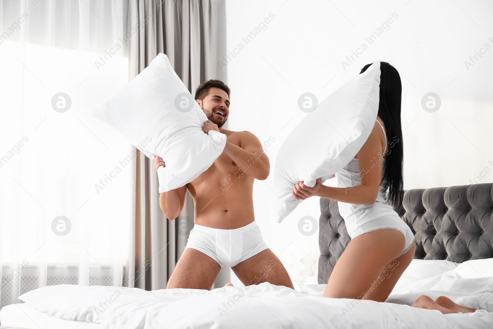 Photo of Young couple in white underwear having pillow fight on bed at home