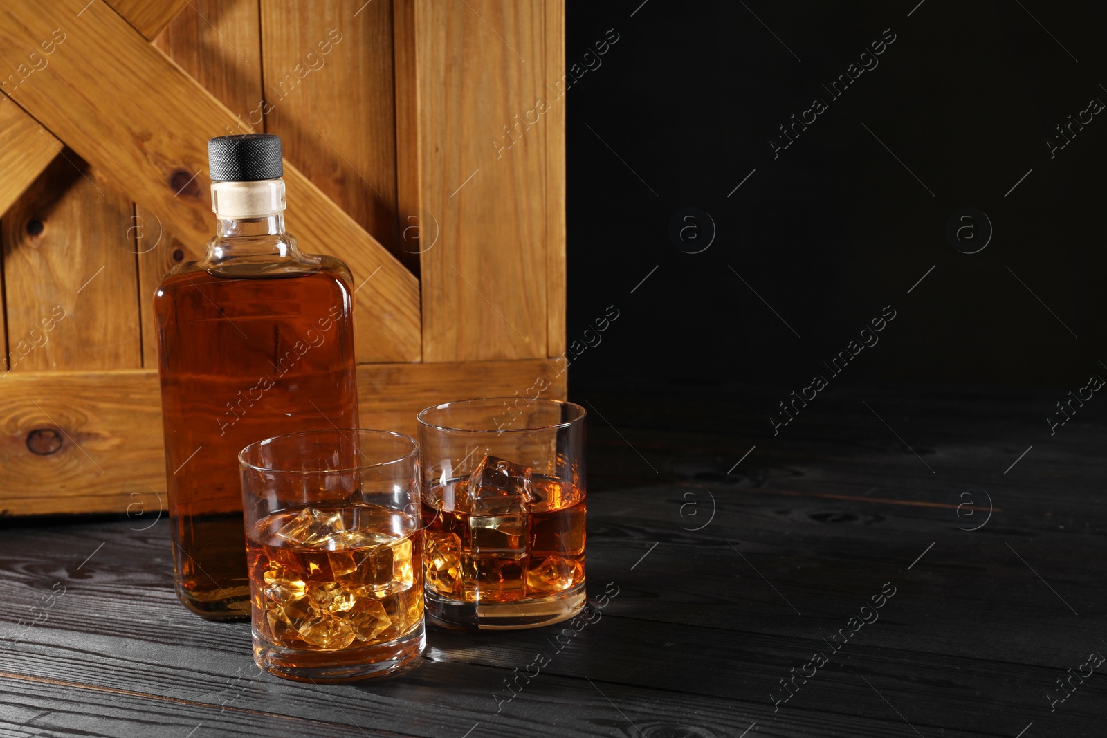 Photo of Whiskey with ice cubes in glasses and bottle on black wooden table, space for text