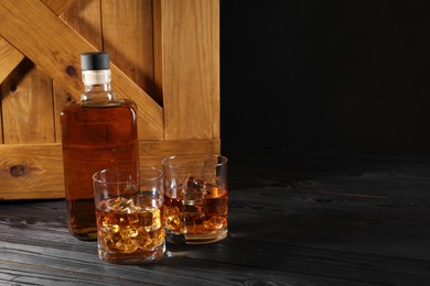 Whiskey with ice cubes in glasses and bottle on black wooden table, space for text