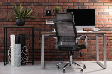 Photo of Cosy workspace with computer on desk, chair and potted plants near brick wall at home