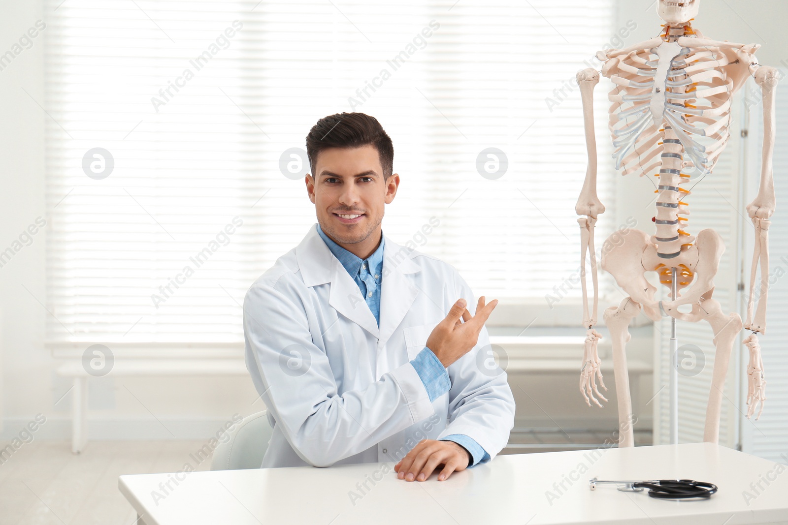 Photo of Male orthopedist at table near human skeleton model in office