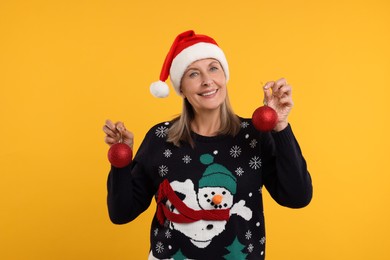 Happy senior woman in Christmas sweater and Santa hat holding festive baubles on orange background