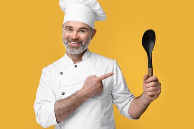 Photo of Happy chef in uniform pointing at spoon on orange background