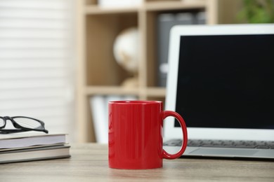 Photo of Red ceramic mug, notebooks and laptop on wooden table indoors