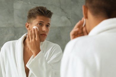 Photo of Handsome young man cleaning face with cotton pad near mirror in bathroom