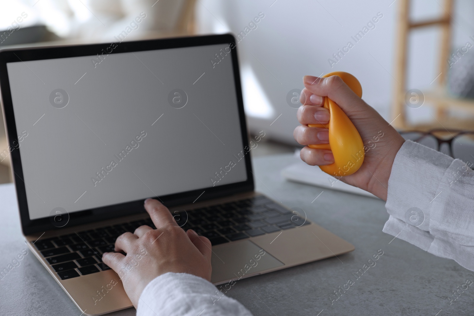 Photo of Woman squeezing antistress ball while working on laptop in office, closeup