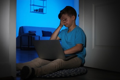 Photo of Frightened little child with laptop on floor in dark room. Danger of internet