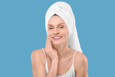 Beautiful woman in terry towel removing makeup with cotton pad on light blue background