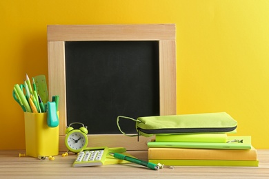 Different school stationery and small blank chalkboard on table near yellow wall. Space for text