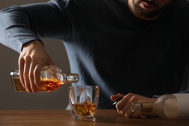 Photo of Addicted man pouring alcoholic drink into glass at wooden table indoors, closeup