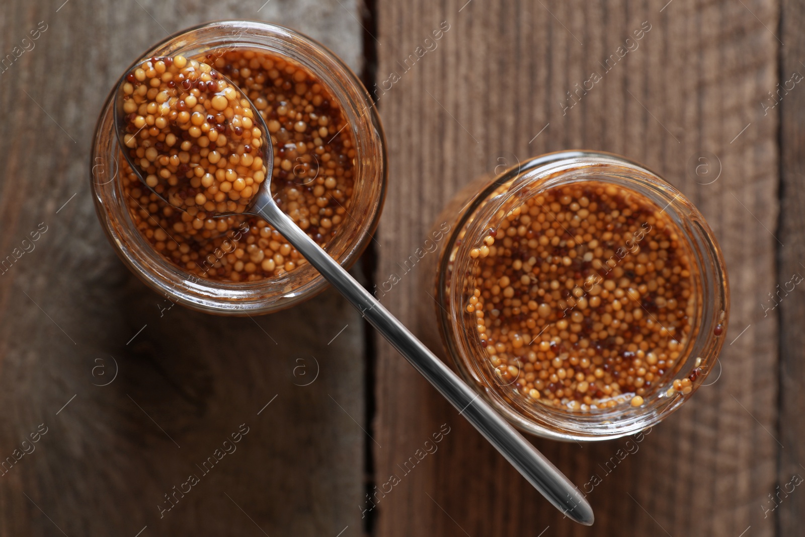 Photo of Jars and spoon of whole grain mustard on wooden table, flat lay