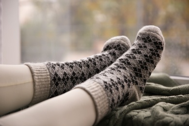 Photo of Woman wearing warm socks on knitted plaid near window, closeup. Cozy season