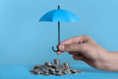 Photo of Woman holding small umbrella over coins on light blue background, closeup
