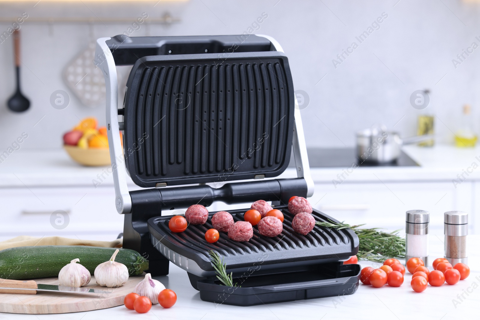 Photo of Electric grill with meatballs, tomatoes and vegetables on white table in kitchen