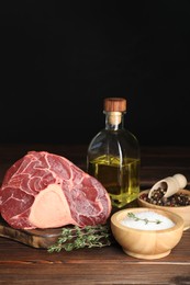 Piece of raw beef meat, thyme, oil and spices on wooden table against black background