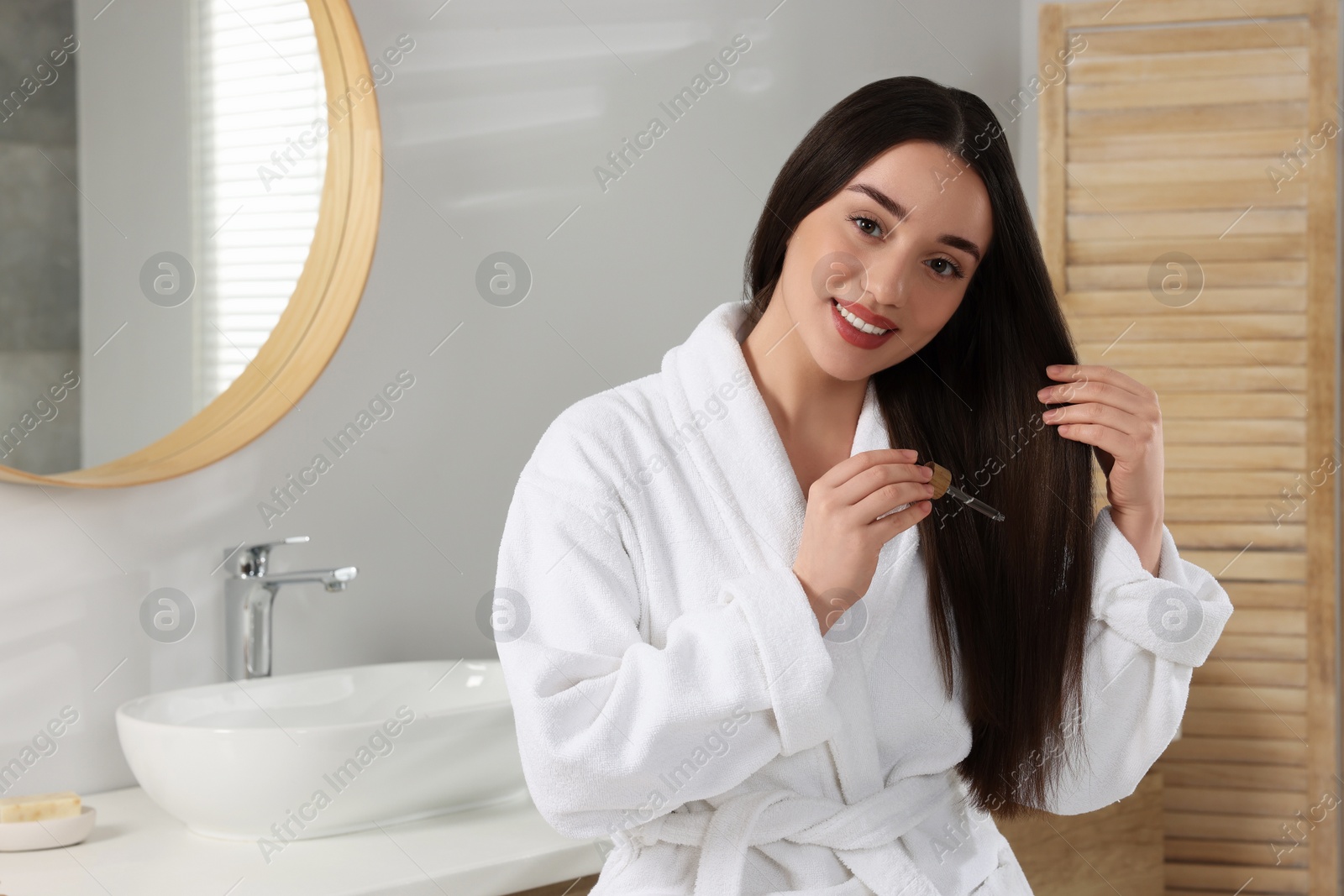 Photo of Happy young woman applying essential oil onto hair in bathroom