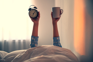Photo of Woman with cup and alarm clock lying in bed, closeup. Morning time
