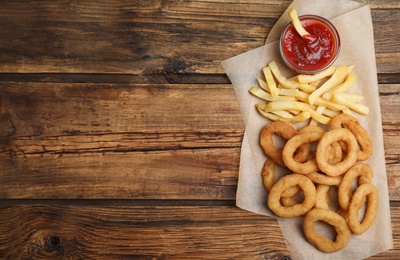 Delicious onion rings, fries and ketchup on wooden table, top view. Space for text