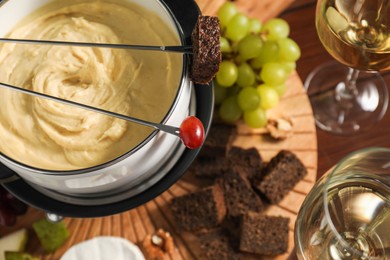 Forks with pieces of grape, bread, fondue pot with melted cheese, wine and snacks on table, above view