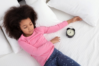 Photo of Cute little African-American girl with alarm clock sleeping in bed, above view
