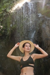 Beautiful young woman in stylish bikini and hat near waterfall outdoors, low angle view
